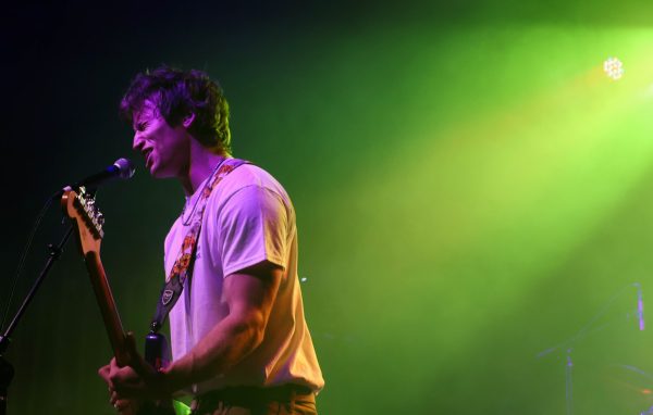 Viewed from the side, a performer squints as he sings passionately into a microphone and plays his electric guitar, a green stage light shining down from behind him.