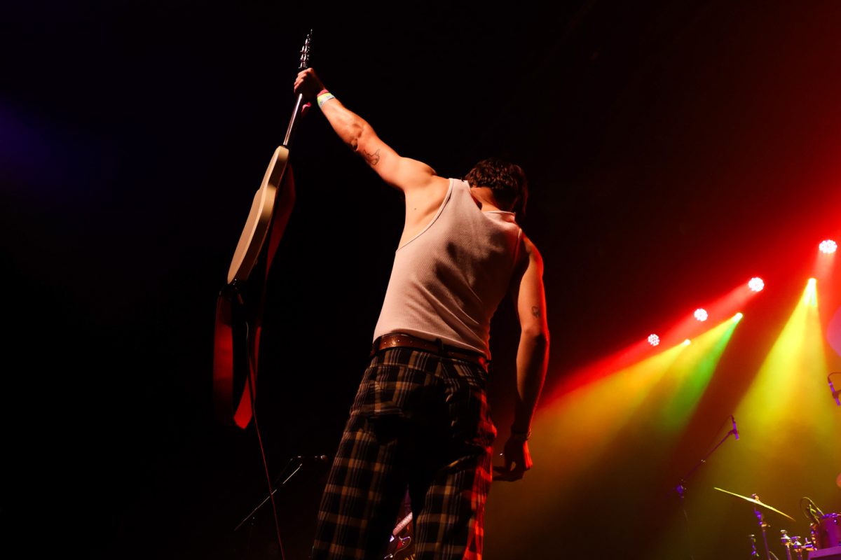 Viewed from behind, a performer looks down while holding his guitar in his left hand, his arm fully extended in a dramatic manner.