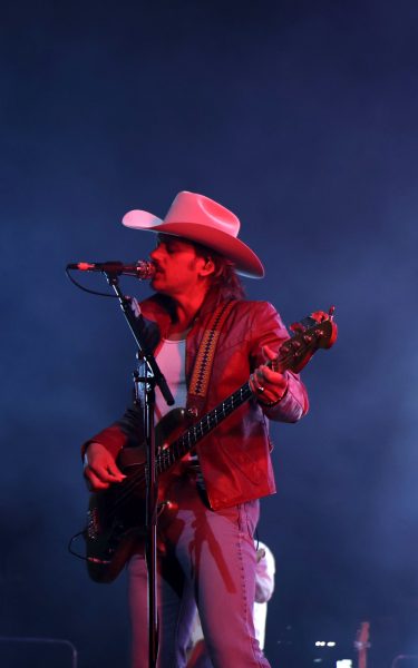 Lit by red stage lights, a country musician in Western wear sings into a microphone while playing his guitar.