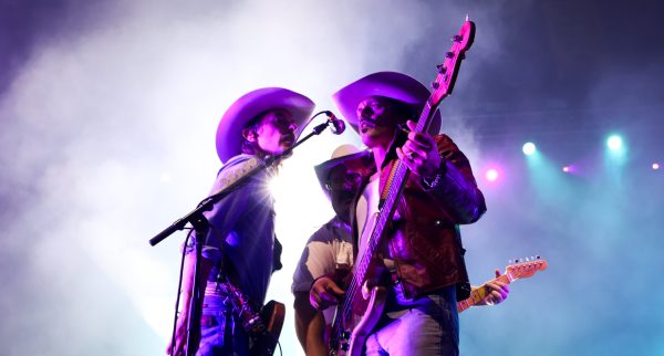 Lit by purple stage lights, three country musicians in Western wear, holding guitars, gather around one microphone.