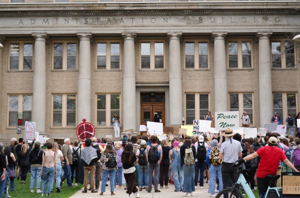 Viewed from behind, a large crowd of protesters stands in front of a building with stone Roman-style columns and many windows.