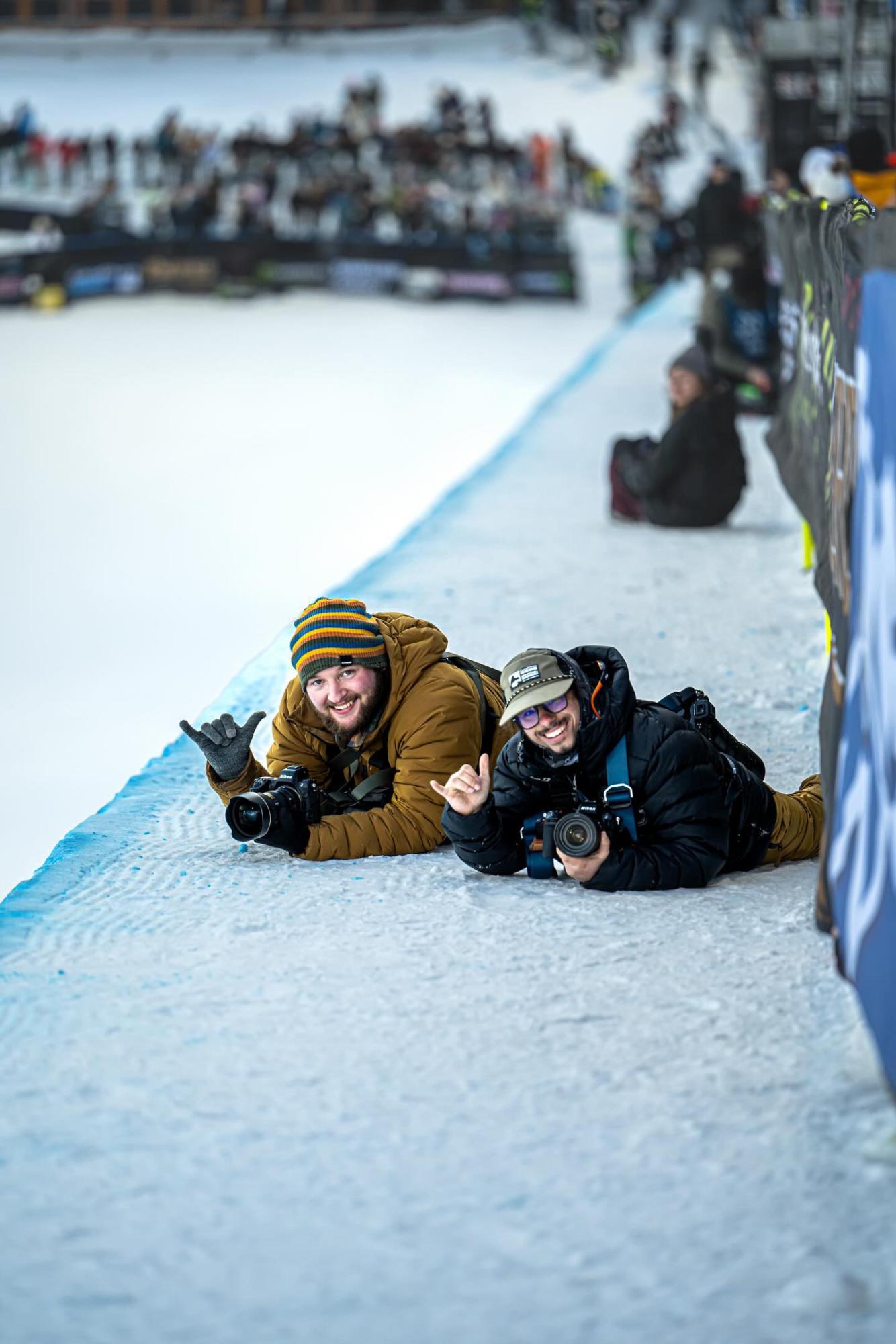 Two photographers lie in the snow with their cameras, both holding up a hang loose sign with their hands.