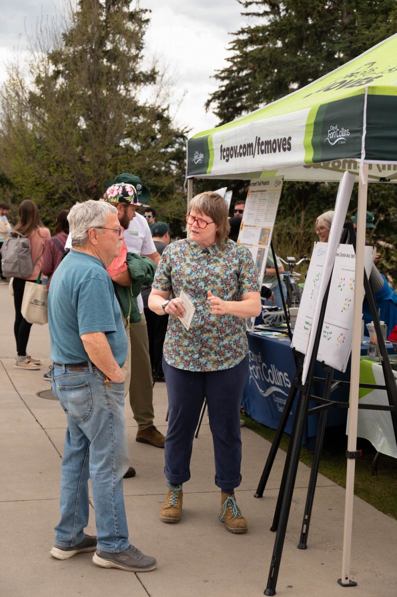 A woman talks to a man next to a table and a pop-up canopy decorated with the City of Fort Collins logo and the URL, fcgov.com/fcmoves.