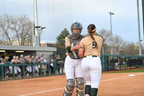Two softball players, one wearing an umpires uniform, lightly embrace while on the softball field.