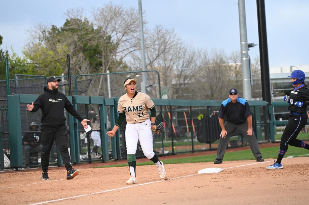 A softball player in a white, gold and green uniform shouts while standing to the left of a base on a softball field. A player in a black and blue uniform runs for the base, and two men focus on the game from the background.
