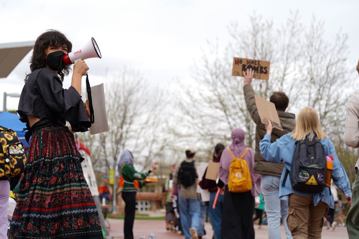 A line of protesters holding signs walks away from the camera on the right while one stands on the left with a megaphone held to their face.
