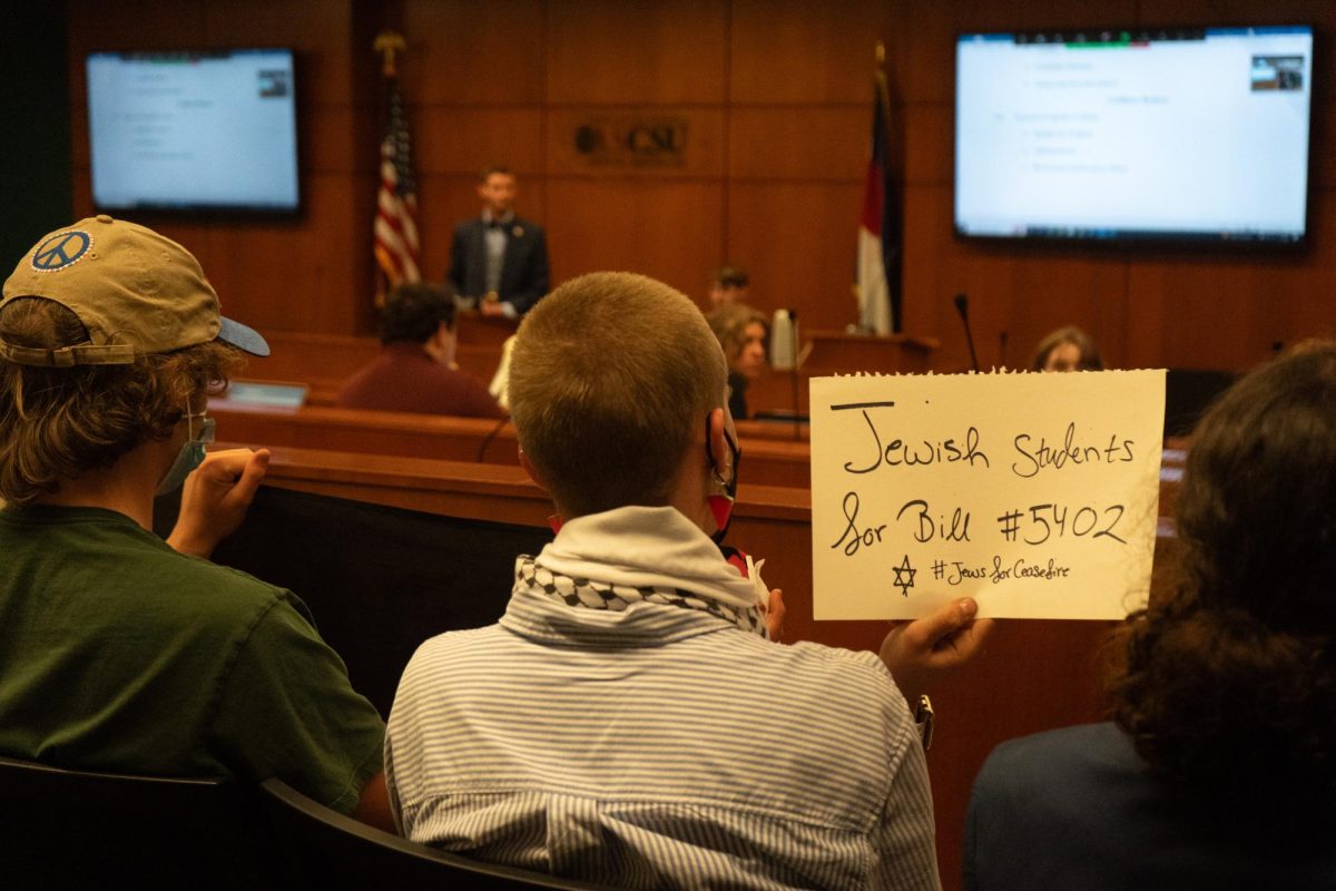 Viewed from behind, three people sit in a row of seats near the back of a wood-paneled presentation room. The person sitting in the middle holds a sign reading, Jewish students for Bill #5402 hashtag Jews for Ceasefire.