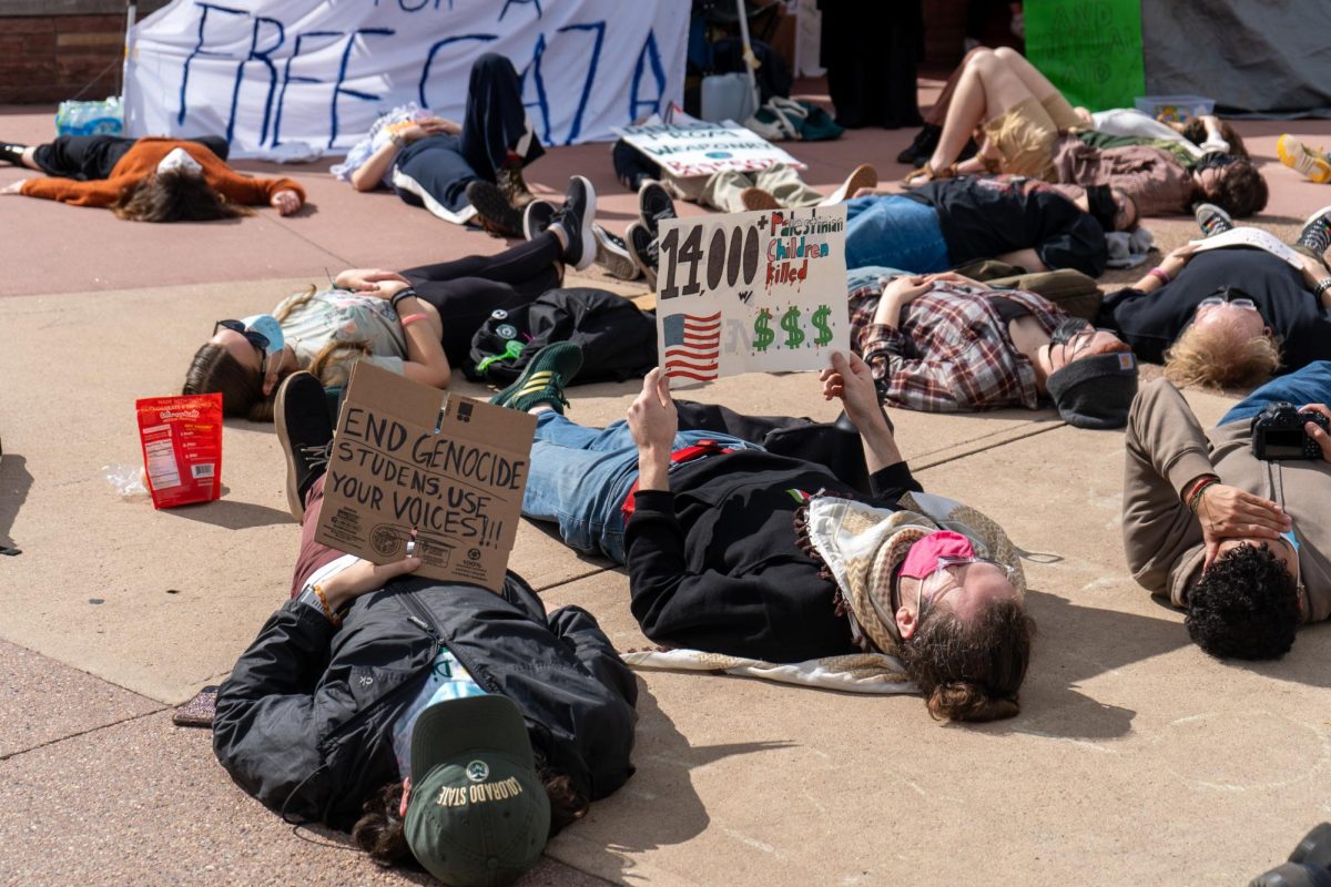 Several protesters lie on the ground while holding up signs. One sign reads, End genocide. Students, use your voices! and another reads, 14,000 plus Palestinian children killed.