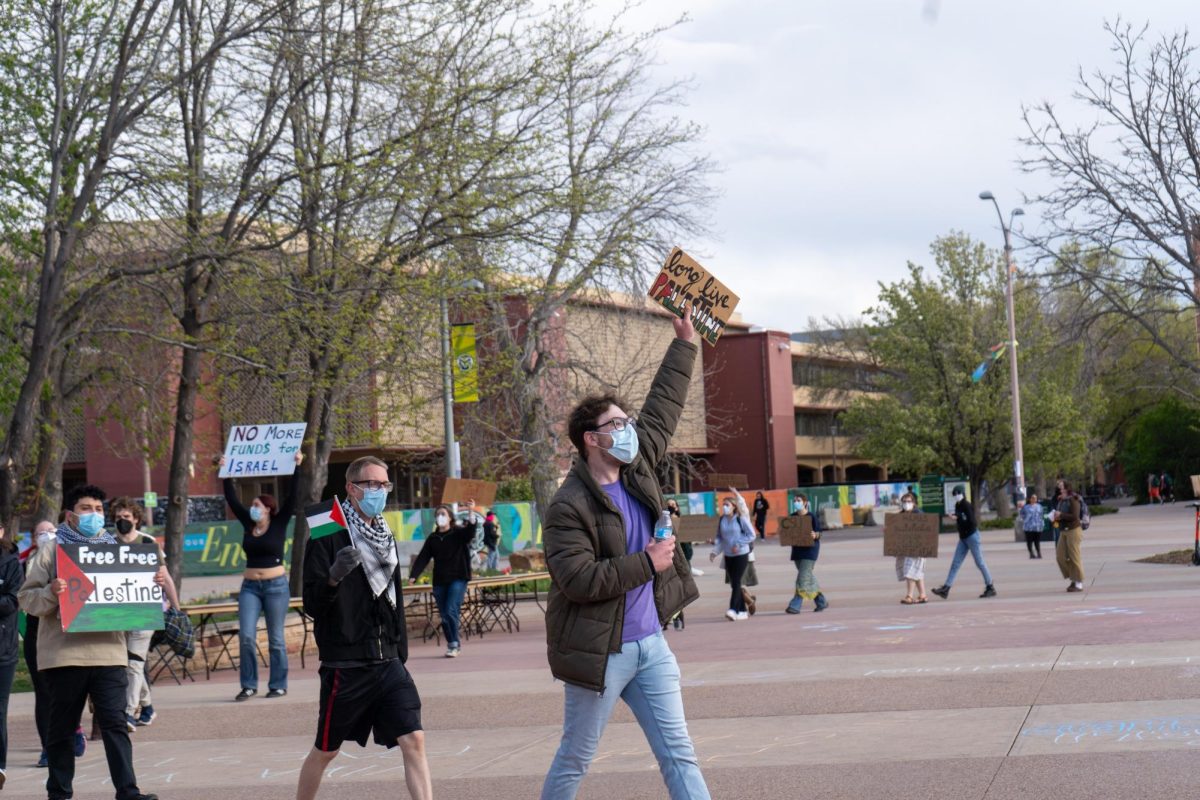 A line of protesters marches on The Plaza with the Andrew G. Clark Building in the background. The protester in the foreground holds a sign above his head reading, Long live Palestine.