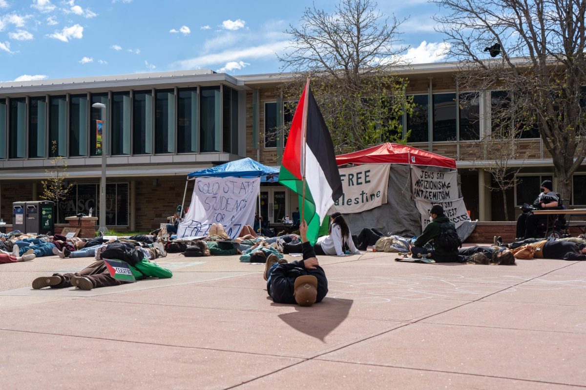 In front of two tent covers and protesters lying on the ground, one protester on the ground holds up a Palestinian flag perpendicular to the ground in the center of the photo.