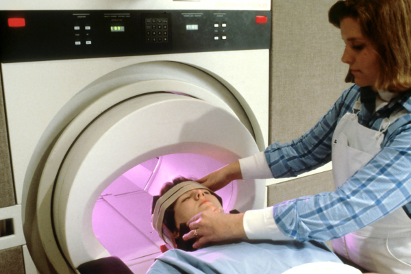An technician calmly positions a patients head as they lie on the patient table of an MRI machine, preparing to enter the chamber of the MRI machine.