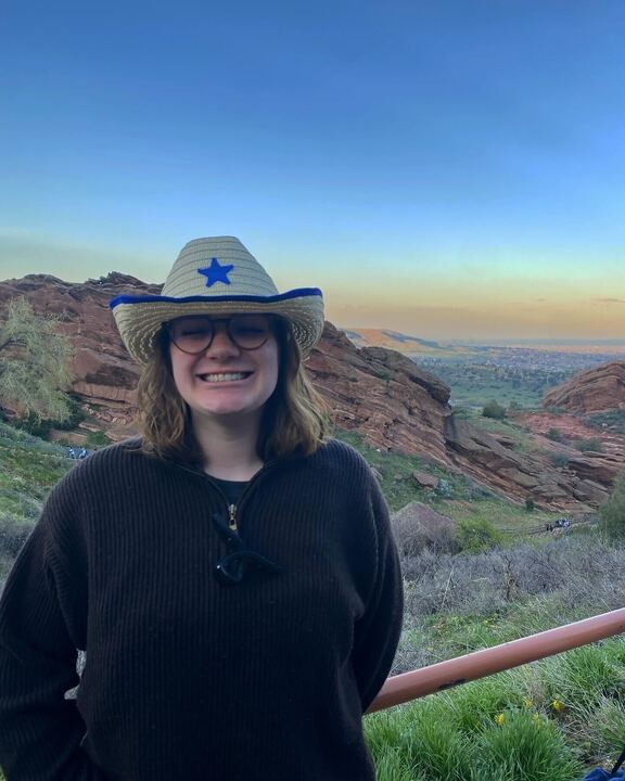 A+woman+wearing+a+cowboy+hat+and+smiling+at+the+camera+stands+in+front+of+a+view+of+red+rocks%2C+shrubbery+and+a+colorful+sky.