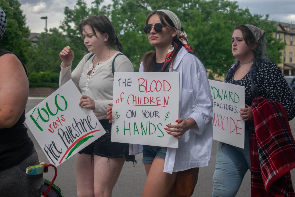 Three protesters holding signs walk down a street. The left sign reads, FoCo says Free Palestine. The middle sign reads, The blood of children on your hands. The right sign reads, Woodward manufactures genocide.