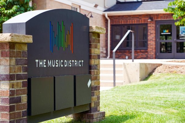 A sign reading, the music district, in front of a short brick building with a lawn and a concrete staircase.