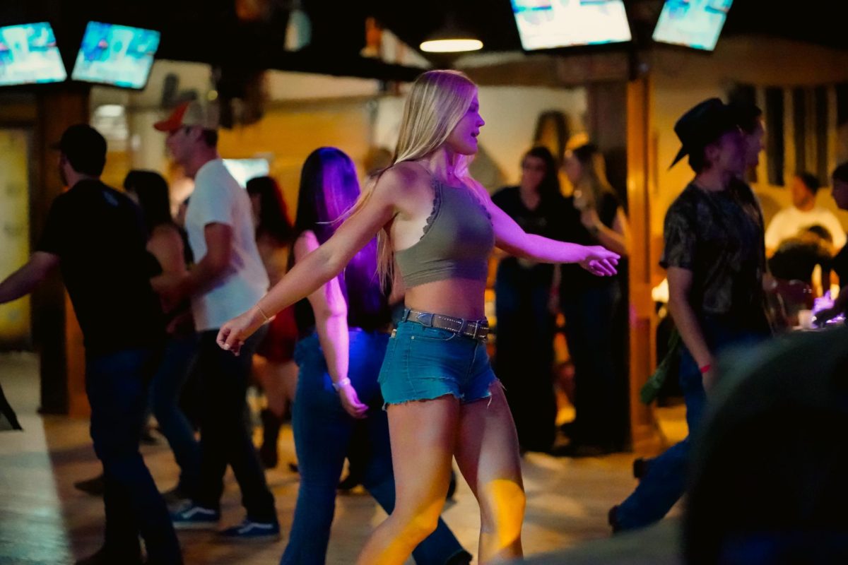 A blond woman in casual clothing dances in the middle of a room full of other dancers.