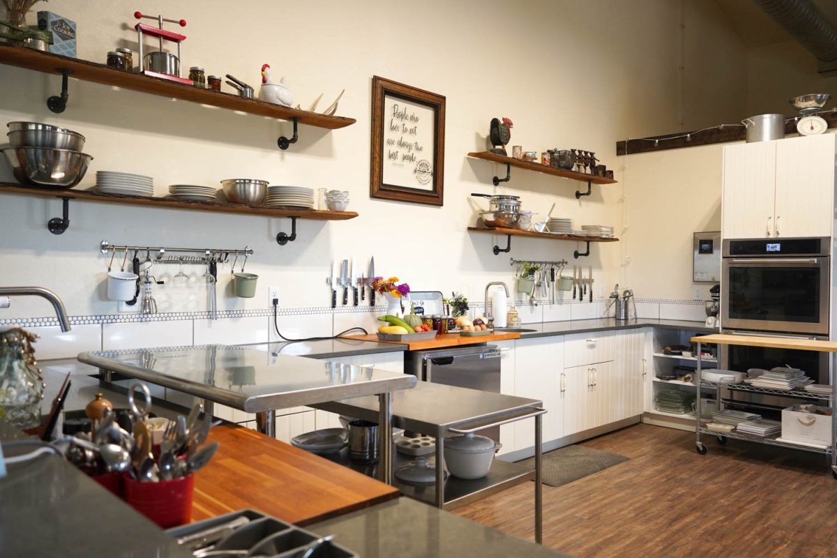 Photo of a kitchen with stainless steel counter tops and white cabinets. There are shelves that have plates and bowls on them. On the counter there are fruits, vegetables, jam, eggs, and a sign that reads, "Don't go Bacon my Heart".