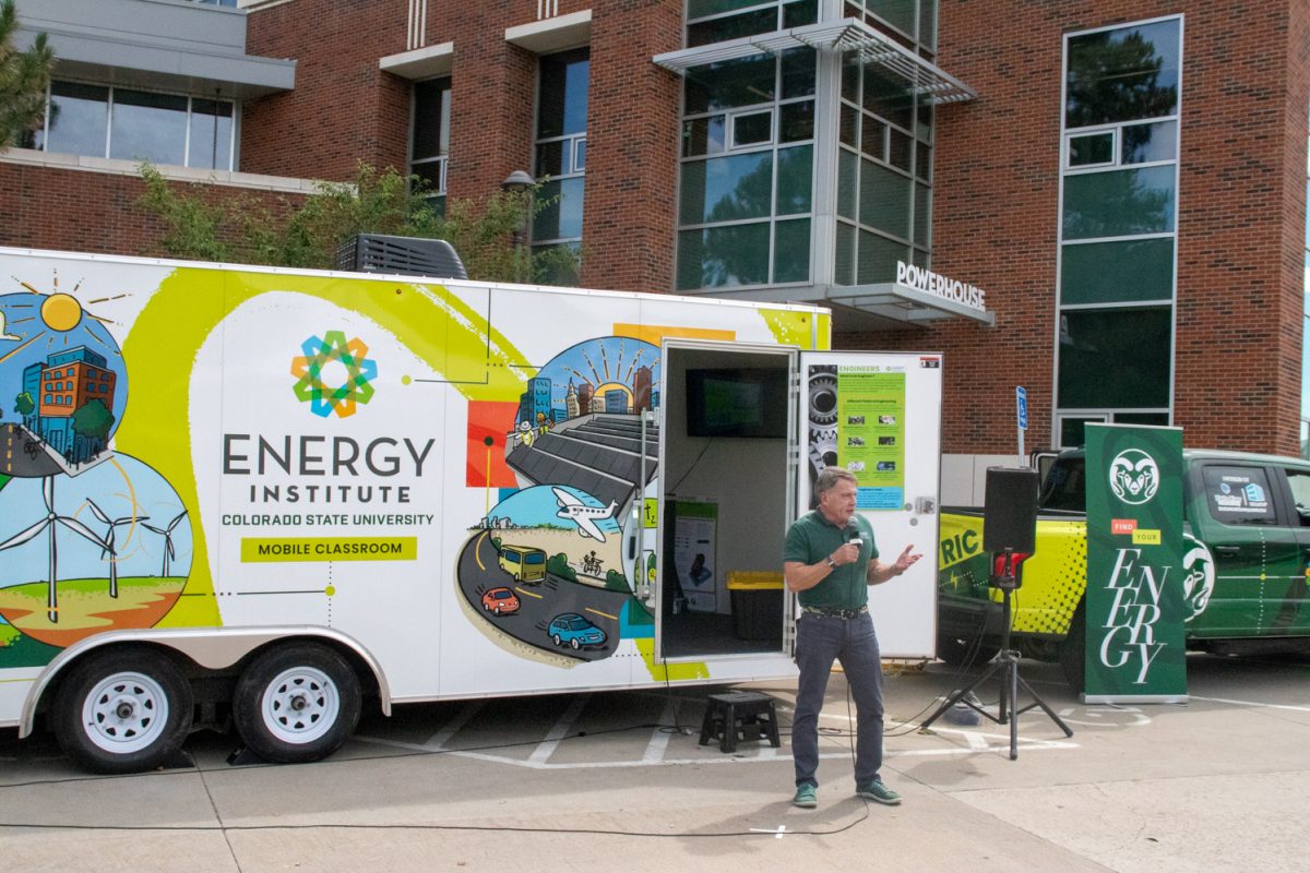 A man stands with a microphone in front of a truck with a trailer that says "Energy Institute Colorado State University Mobile Classroom" with colorful energy related designs on it.
