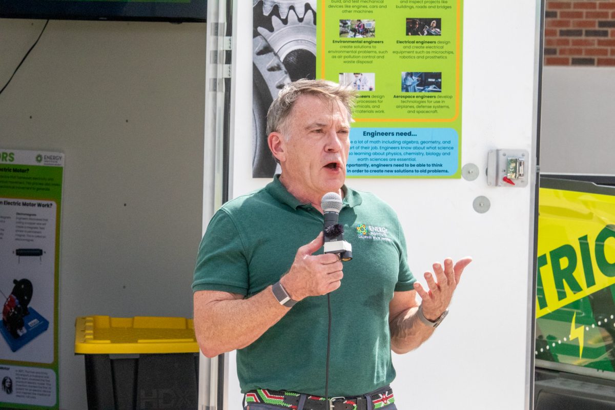 A man stands with a microphone in front of a truck with a trailer that says "Energy Institute Colorado State University Mobile Classroom" with colorful energy related designs on it.