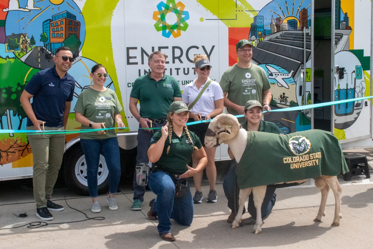 Five people stand preparing to cut a ribbon and two people kneel with a Rambouillet sheep in front of a trailer housing the Energy Institute Mobile Classroom.