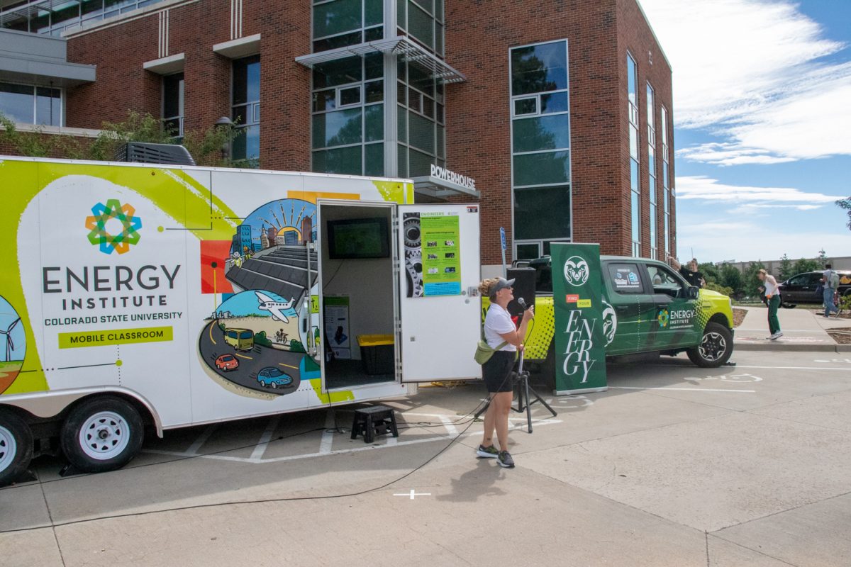 A woman talking into a microphone in front of a truck and a trailer which are parked in front of a building.