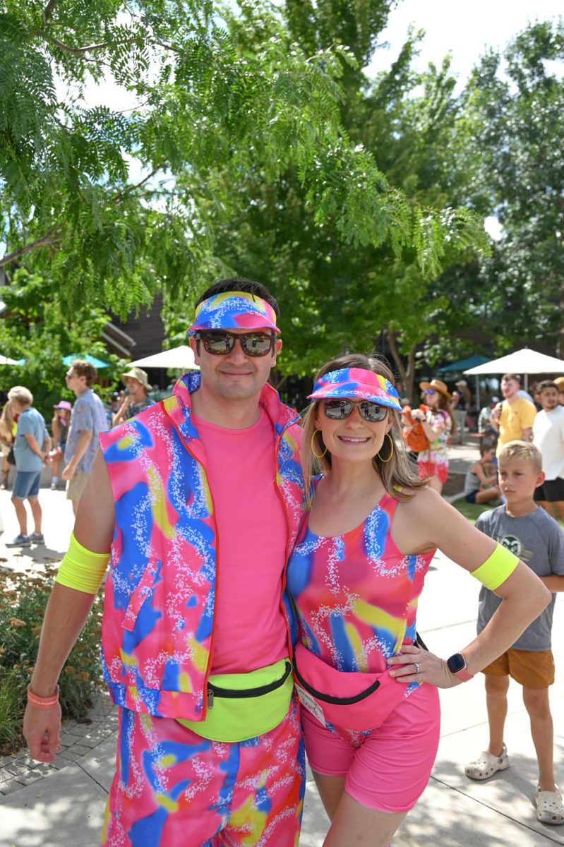 A man and A woman stand outside dressed in a neon blue, pink, and yellow 80's style rollerskating costume with fanny packs and visors to match.