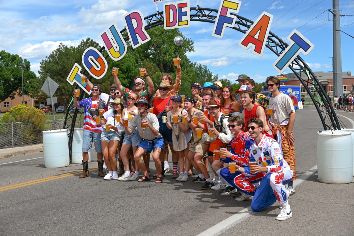 A large group of friends dressed in colorful outfits holds cups of beer while posing in front of an arch that reads "Tour de Fat" in rainbow lettering.