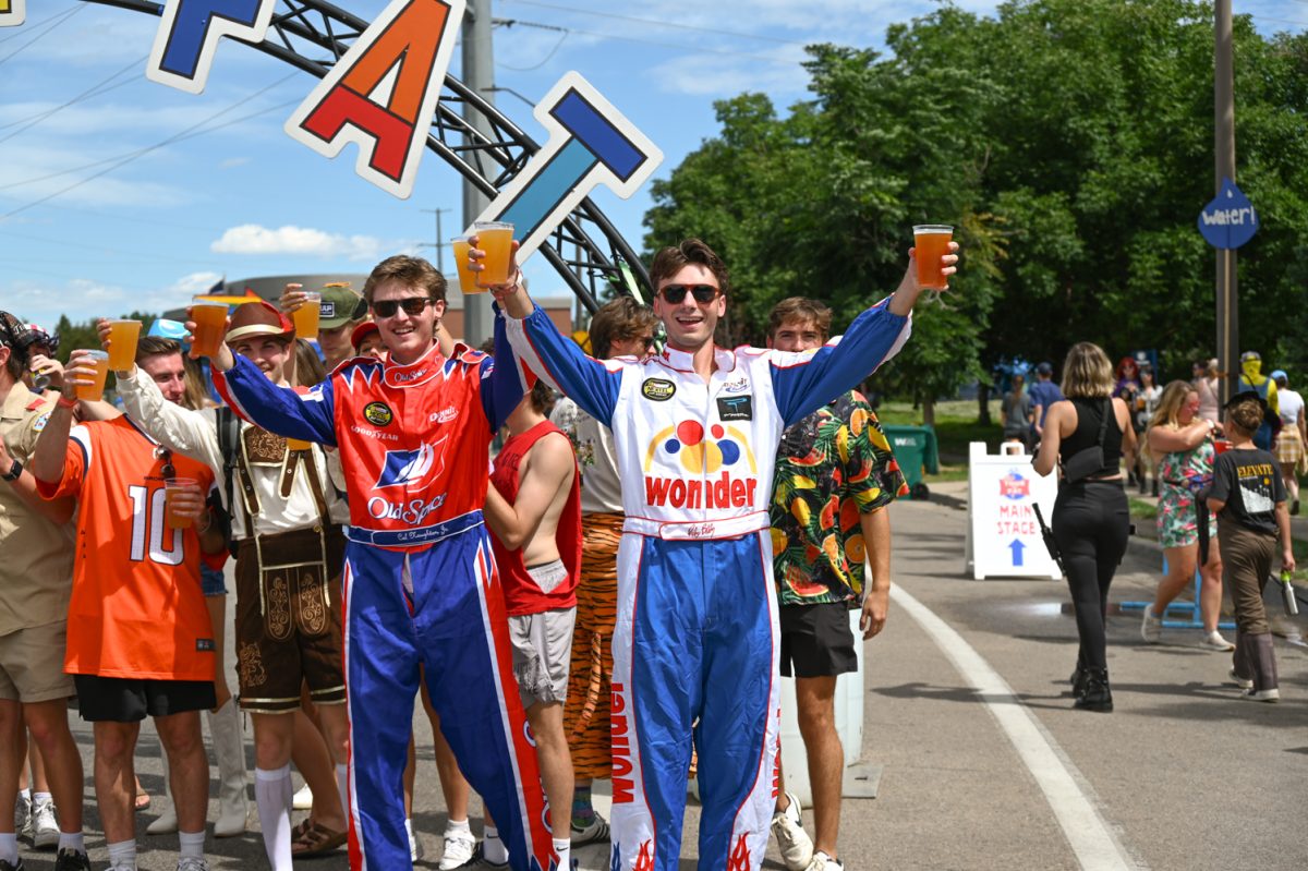 Two men in racing outfits stand in front of a crowd while holding glasses of beers above their heads.