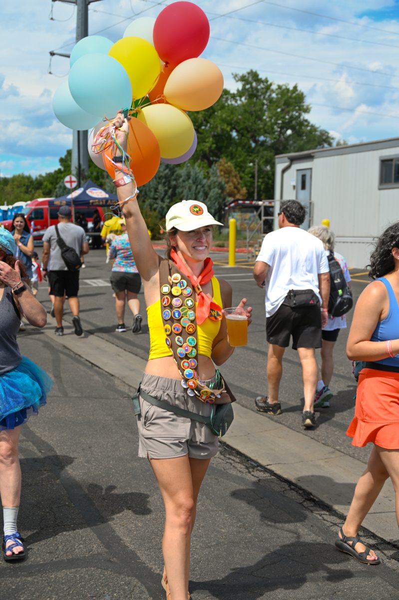 A woman wearing a yellow tank top and a boy scout sash covered in patches walks down a street, one hand holding a glass of beer and the other holding a bouquet of balloons above her head.