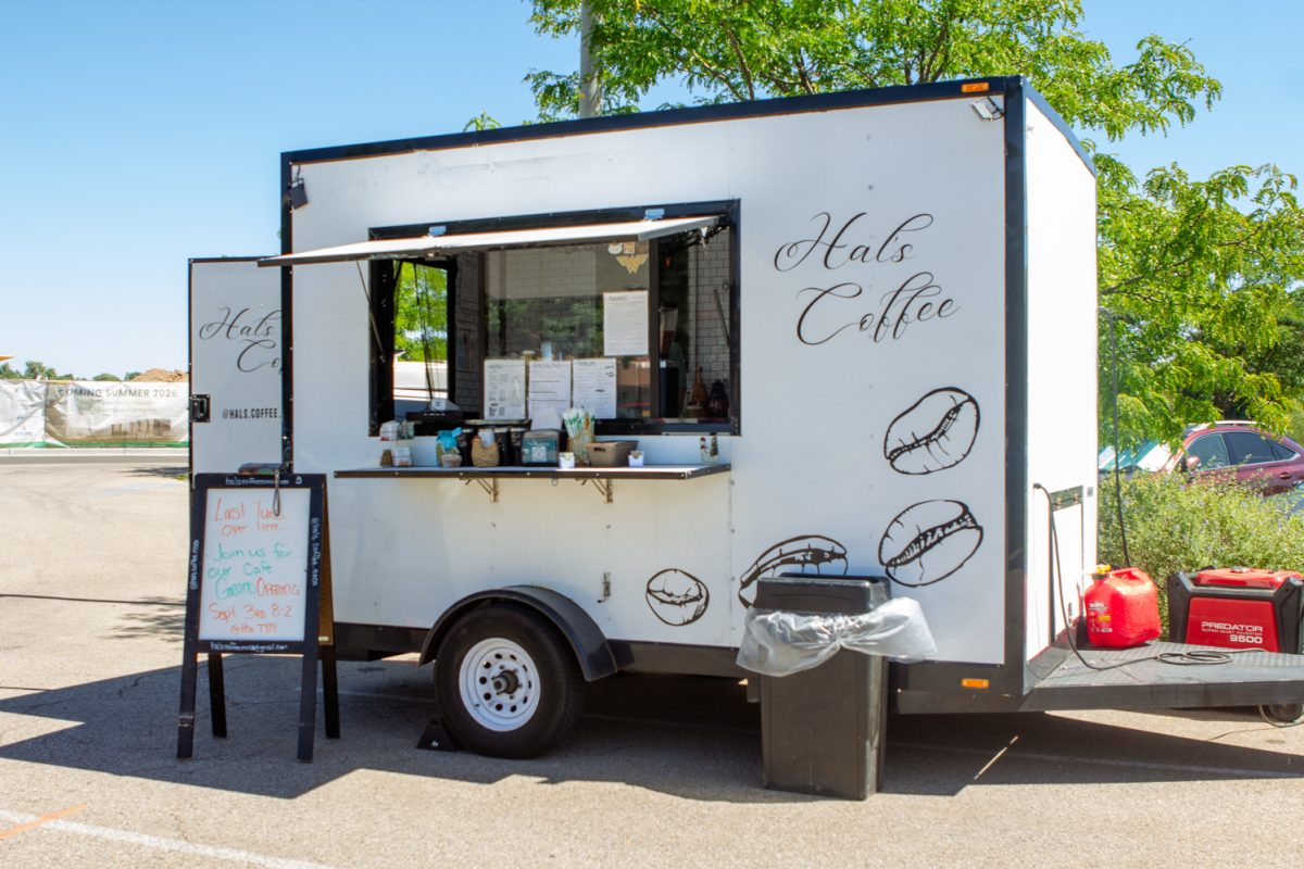 A black and white coffee truck is parked in a parking lot; a white board with writing and a trash can sit outside of the truck. There's a small counter containing merchandise and coffee condiments. Cursive handwriting reads "Hal's Coffee" on the side of the truck.