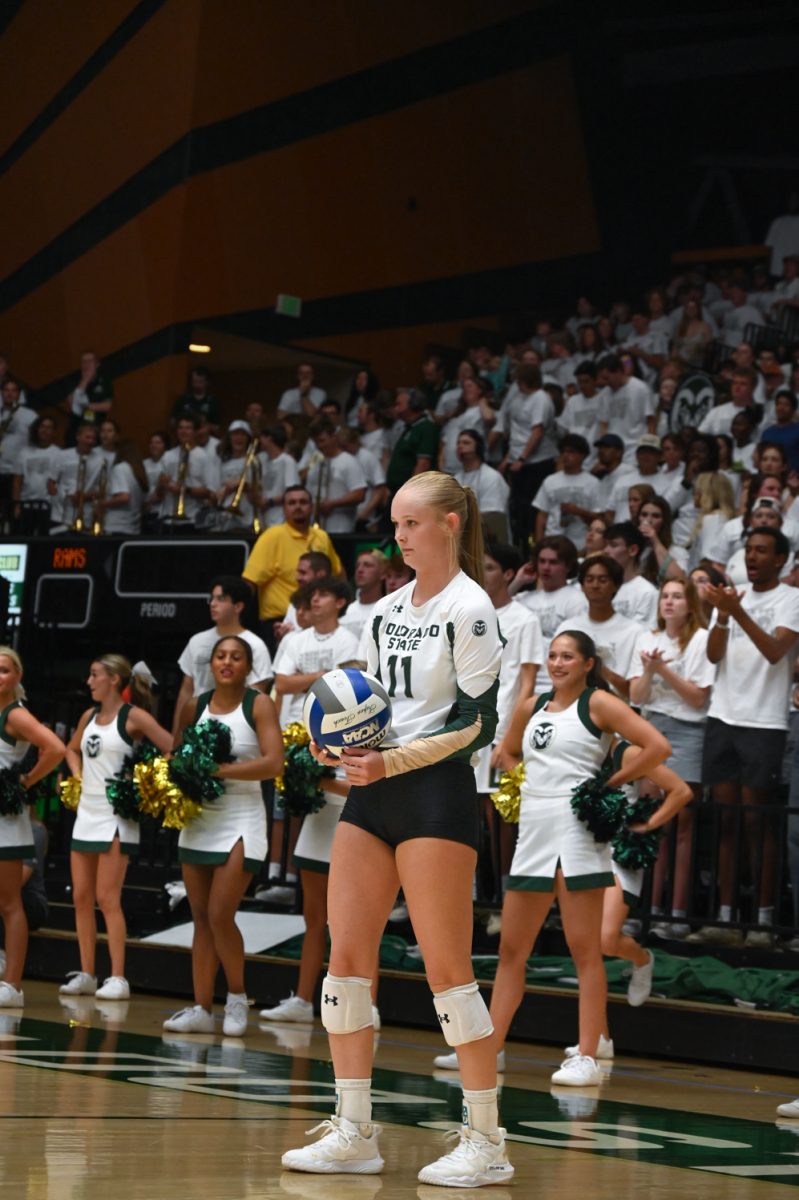 A Girl in a green and gold long sleeve uniform holds a blue and white volleyball on the court in front of a large crowd