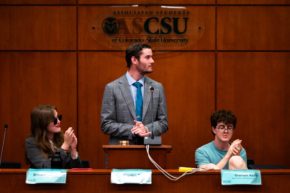 Speaker of the Senate Hayden Taylor stands at a speaking podium looking to his left and clapping. To his left sits Speaker Pro Tempore Brooke Reese in sunglasses and to his right sits Recruitment and Retention Officer Graham Kelly, both clapping as well. Behind them is a clear sign with the ASCSU logo spelled out.