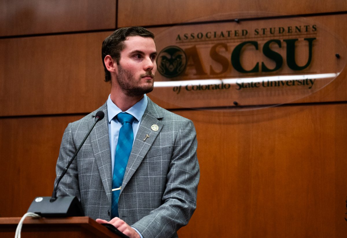 Speaker of the Senate Hayden Taylor stands at a speaking podium and looks to the left. Behind him is a dark-stained wood wall and a sign with the logo of Associated Students of Colorado State University (ASCSU).