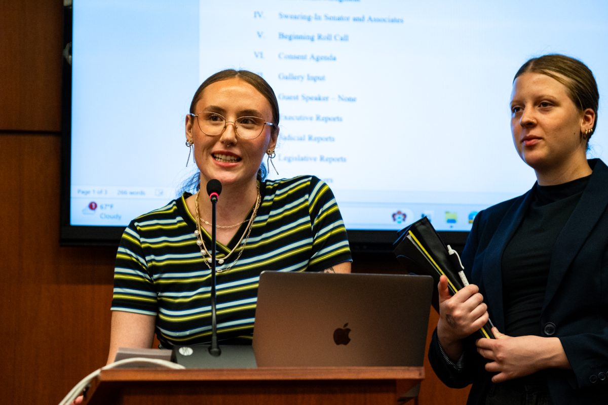 A woman in a striped shirt talks at a podium with a laptop with another woman holding a folder to the right.