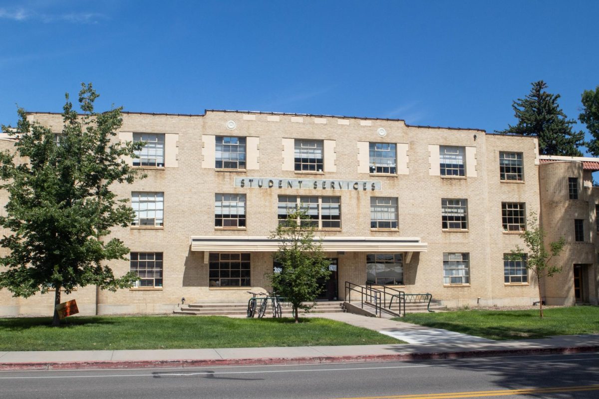 A three story building stands on green grass with two trees on either side. The building is old brick with some brown aged marks. There are two bike racks on either side of the entrance, the rack on the left side has three bikes locked onto it.