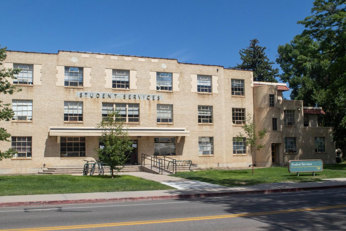An old three story brick building stands on a sunny day on green grass. There is a green Colorado State University building sign on the right marking the building and it's name.