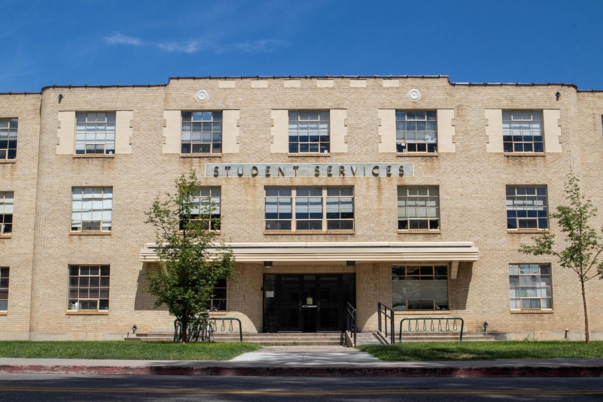 A three story tall aging brick building stands atop green grass with two bike racks, one on each side of the entrance.