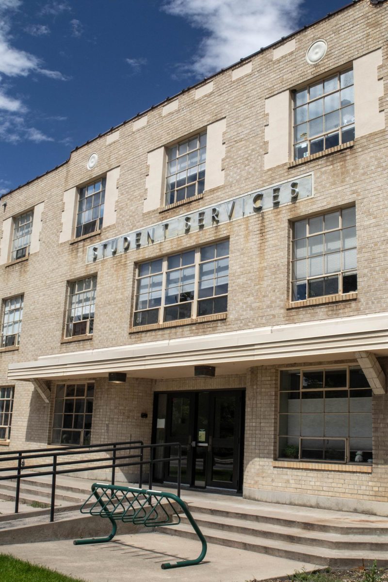 A side image of a three story tall brick building. An empty green bike rack is next to the entrance.