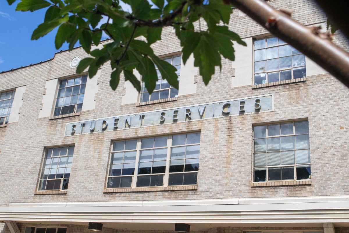 An old brick building stands with letters spelling Student Services across the front of the building. The building is viewed through a tree branch and some leaves
