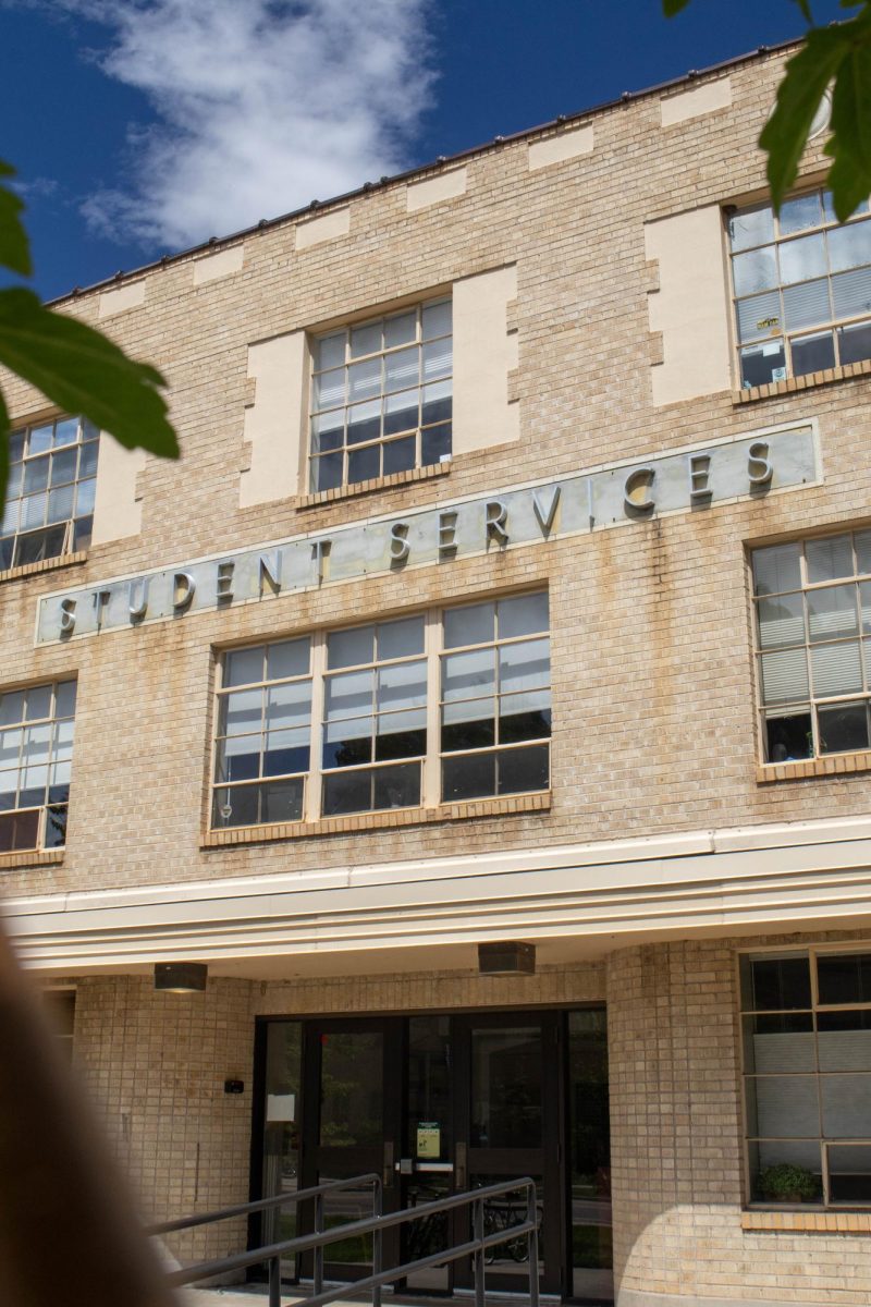 A three story old brick building stands on a sunny day with the words Student Services across the front of the building above the entrance. The image is seen through a few leaves, with the building as the main focus.