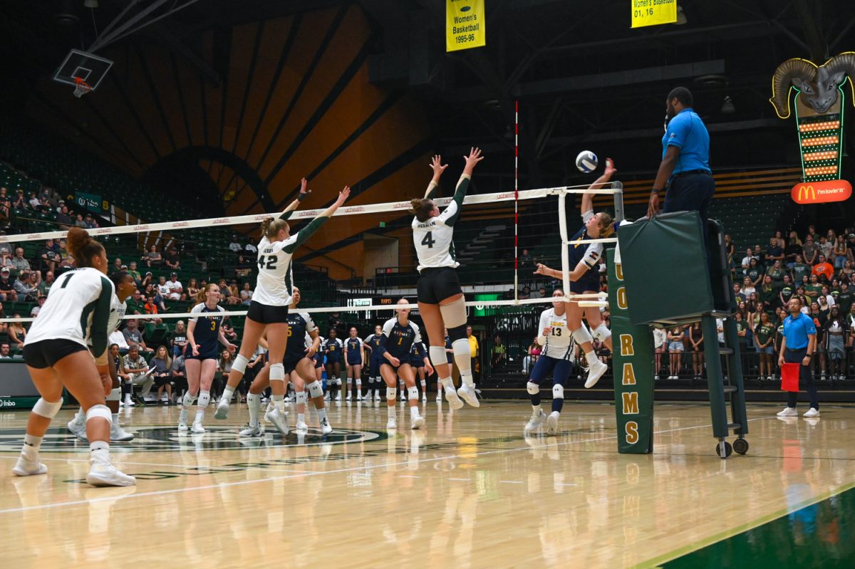 two girls dressed in green and white long sleeve uniforms with tight black shorts jump at the net to block a blue and white volleyball