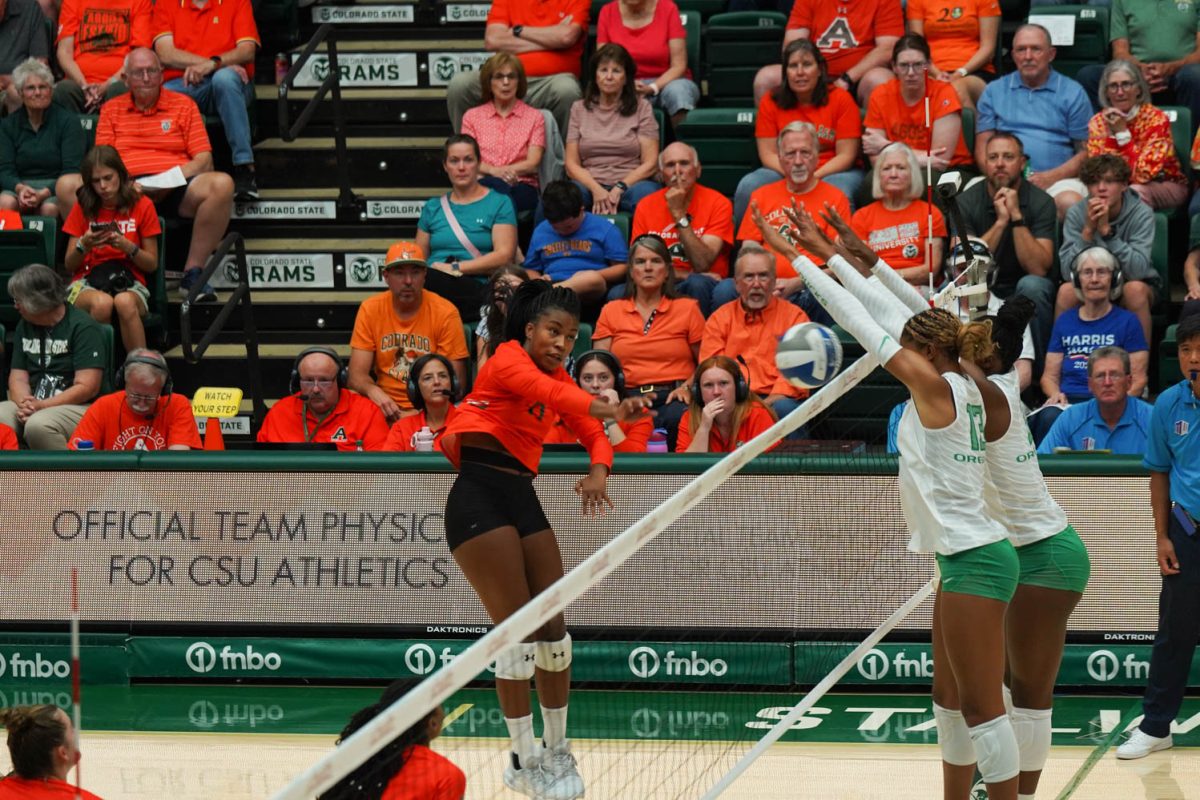 A woman in an orange jersey jumps up to a volleyball net with her hand extended while two women in white and green jerseys on the other side jump up with their hands extended.