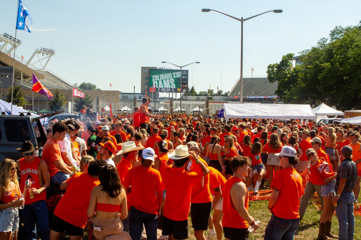 A crowd of people wearing orange clothing mingle around cars and tents outside of a football stadium.