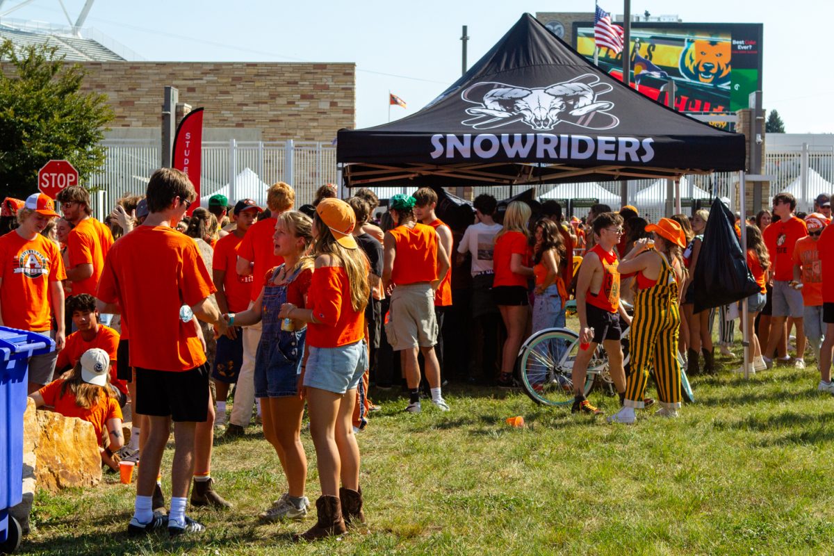 People wearing orange and yellow clothing crowd under and around a tent labeled "SNOWRIDERS".