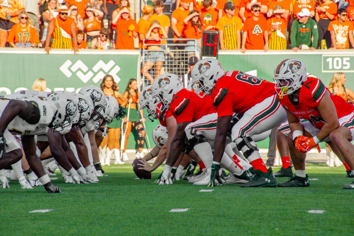 Football players wearing bright orange uniforms crouch and face football players wearing white uniforms as they prepare to start a play on a football field.