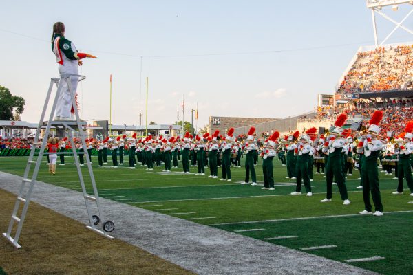 A marching band wearing green and bright orange is lined up on a football field, while a conductor conducts from a raised platform.