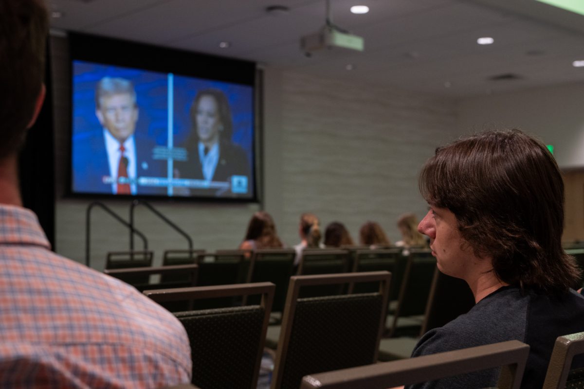 A side profile of a man sitting in a room of chairs watching a projection of the presidential debate between a man in a blue suit and red tie and a woman in a black suit jacket and white shirt.