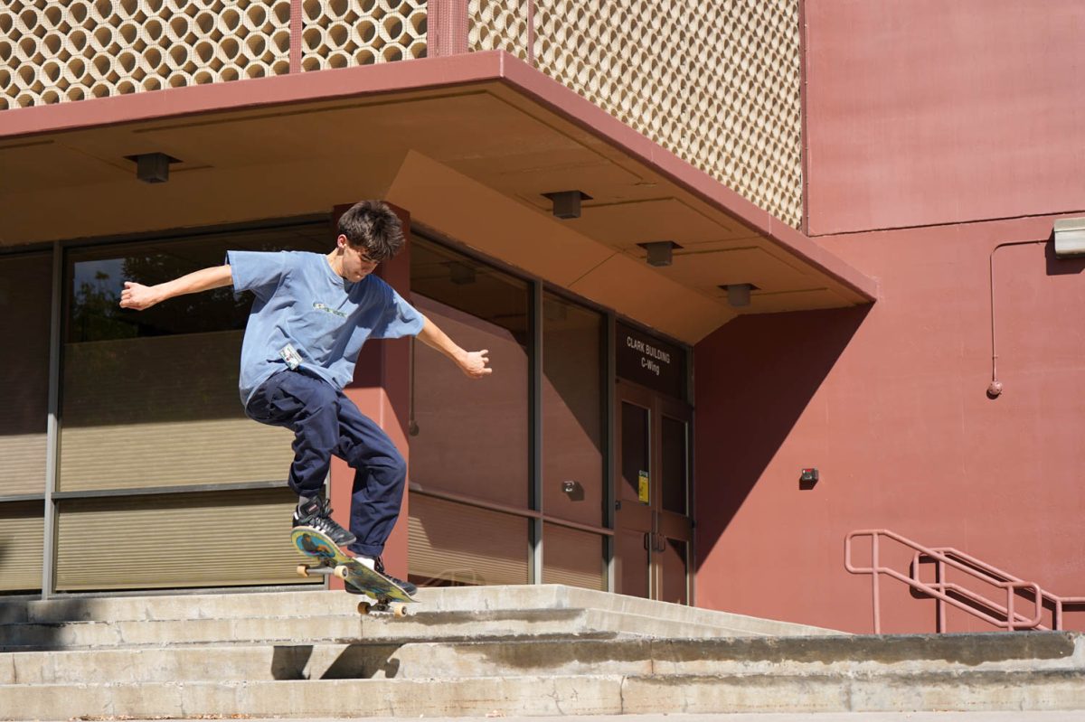 A man in a blue shirt and blue pants skateboards on the steps in front of a red and tan building.