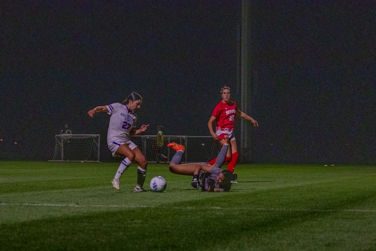 A soccer player is kicking the ball while a goalie is on the ground trying to block her.