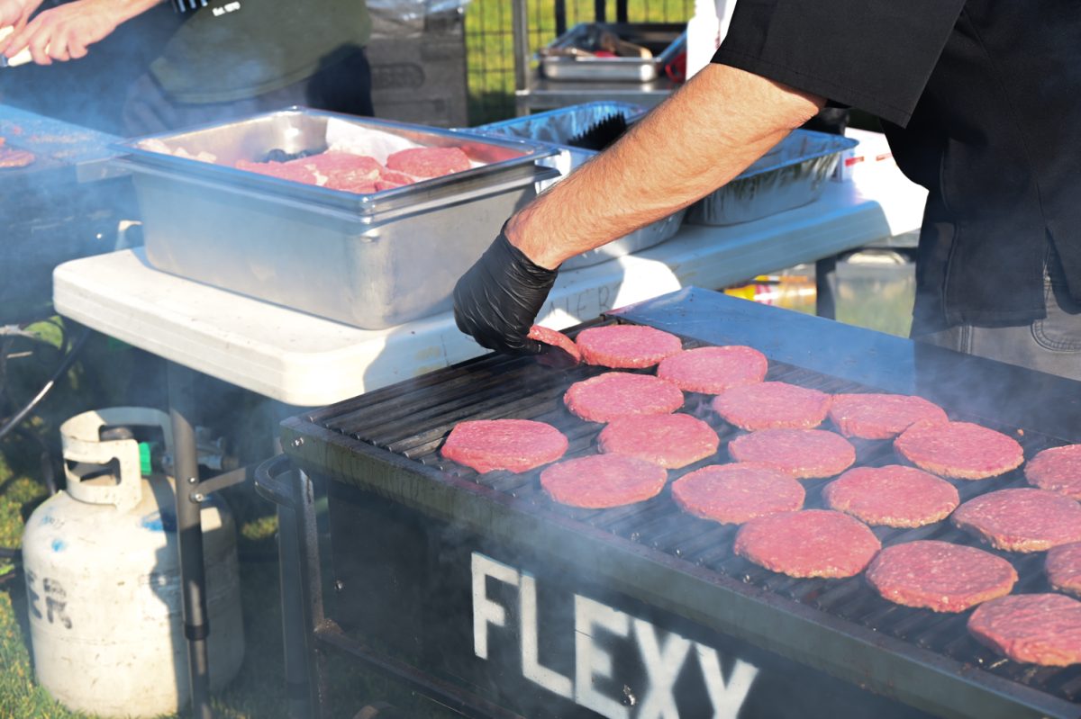 A gloved hand flips one of many meat patties on a steaming grill.