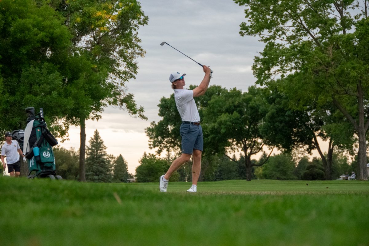 Man in white shirt and hat uses golf club to hit golf ball, sky and trees in background